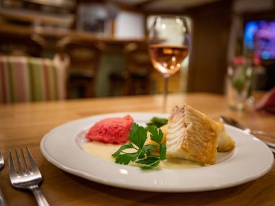 Gebratenes Fischfilet mit roter Beilage und grüner Petersilie auf weißem Teller, Glas Wein im Hintergrund