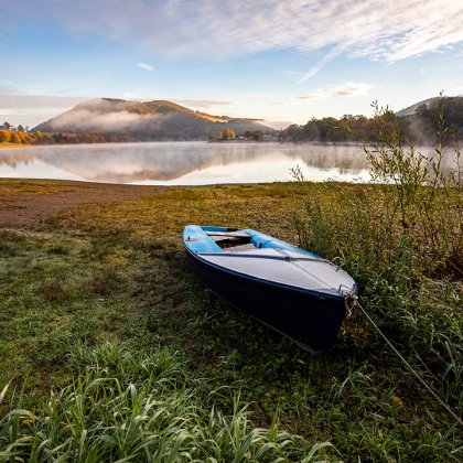 Ein Boot liegt am Ufer eines ruhigen Sees mit Nebel und bewaldeten Hügeln im Hintergrund
