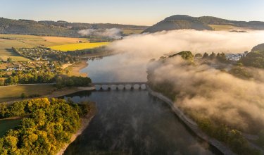 Landschaft mit Fluss, Brücke und Nebel, der über bewaldete Hügel zieht