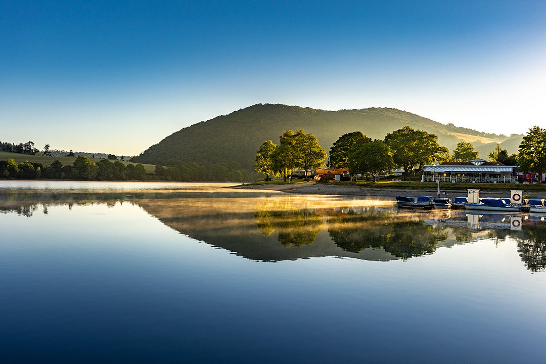 Spiegelglatter See mit Nebel, Bäumen und Hügeln im Hintergrund bei Sonnenaufgang