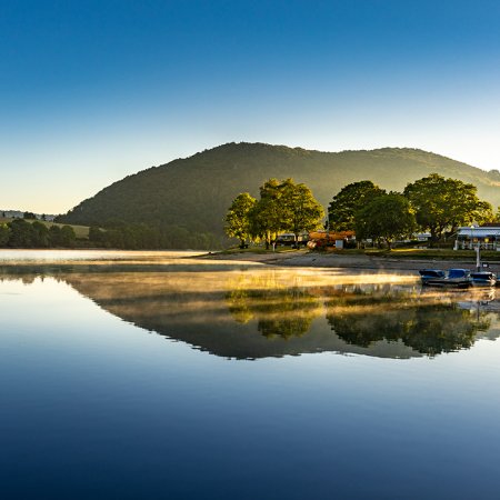 Spiegelglatter See mit Nebel, Bäumen und Hügeln im Hintergrund bei Sonnenaufgang