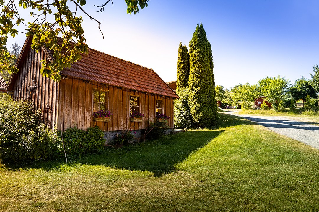 Holzhaus mit roten Dachziegeln links, hohe Zypressen daneben und Weg rechts bei sonnigem Himmel