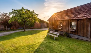 Sonnenuntergang hinter einem Holzhaus mit Bank auf grünem Rasen und Baum links