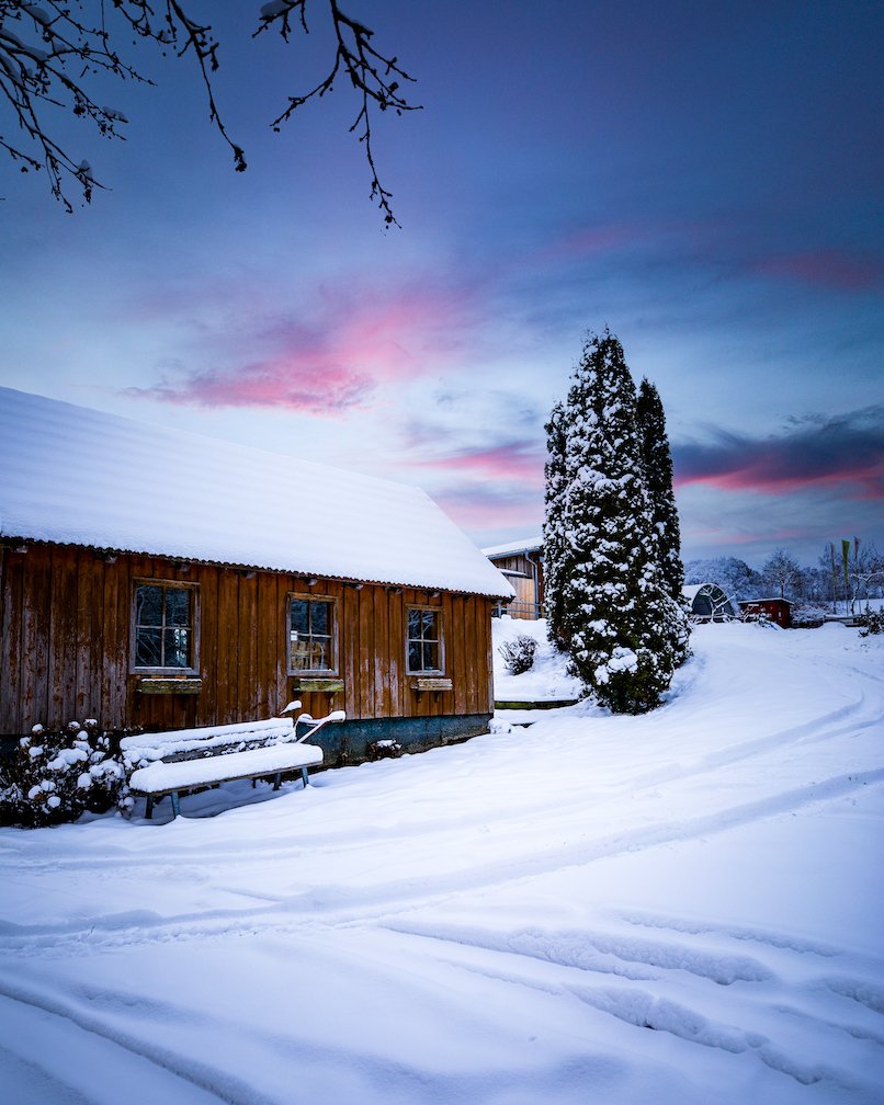 Holzhaus mit schneebedecktem Dach und Bank im Vordergrund, verschneiter Baum rechts, Abendhimmel
