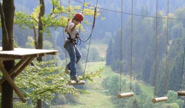 Person mit Helm und Kletterausrüstung balanciert auf hängenden Holzstämmen im Hochseilgarten über Waldtal