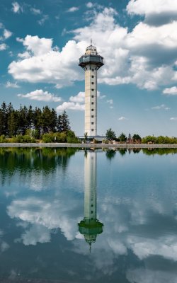 Hoher Aussichtsturm vor bewaldetem Ufer, mit Spiegelung im ruhigen Wasser unter bewölktem Himmel