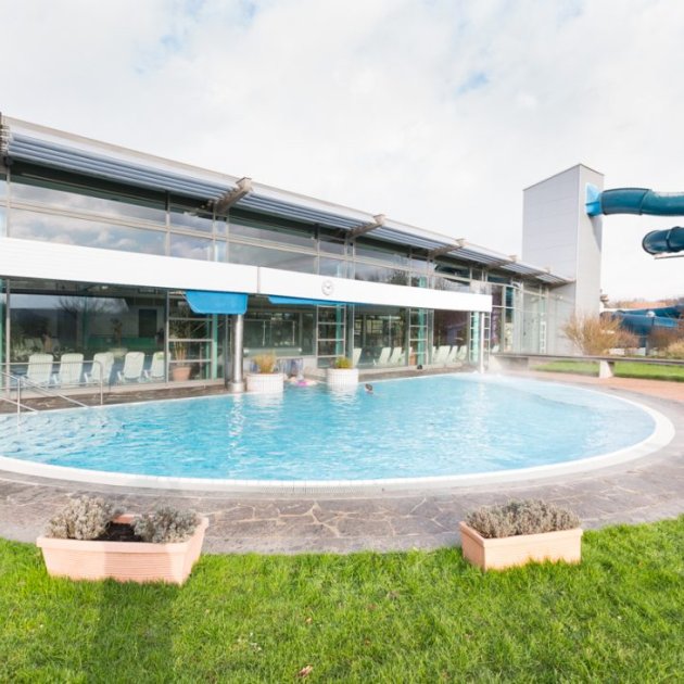 Rundes Außenbecken vor einem modernen Gebäude mit blauer Wasserrutsche rechts und Pflanzen im Vordergrund