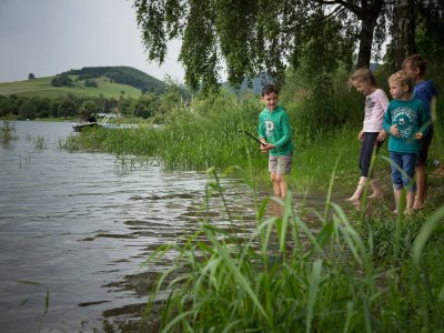 Kinder spielen am Ufer eines Sees mit grüner Vegetation und Boot im Hintergrund