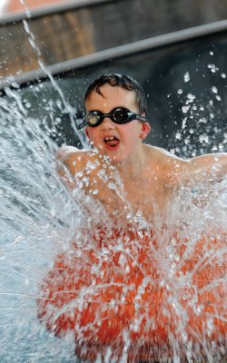 Junge mit Schwimmbrille sitzt in einem Wasserbecken und spritzt Wasser nach oben