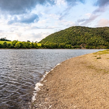 Ufer mit Kiesstrand rechts und bewaldetem Hügel im Hintergrund unter bewölktem Himmel