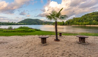 Palme und Holztische auf Sandstrand am See mit bewaldeten Hügeln und bewölktem Himmel