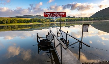 Steg mit Schild 'Seerundfahrten Fährfahrten' über ruhigem See mit Spiegelung von Wolken und Landschaft
