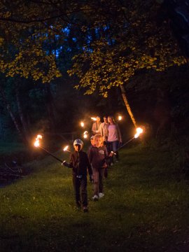 Gruppe von Menschen mit Fackeln wandert nachts auf einem grasbewachsenen Weg im Wald