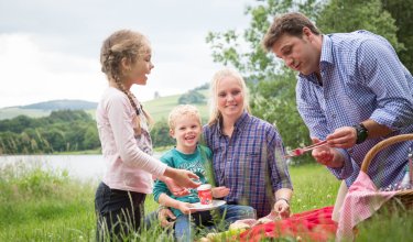 Familie macht Picknick auf einer Wiese am See, Vater hält Gabel, Mutter und Kinder sitzen auf Decke