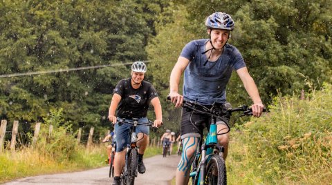 Mehrere Radfahrer fahren auf einer Landstraße bei leichtem Regen, umgeben von Bäumen und Gras