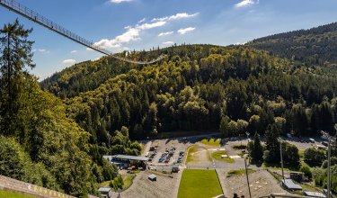 Hängebrücke über bewaldeten Hügeln mit Parkplatz und Sportanlage im Tal unter blauem Himmel