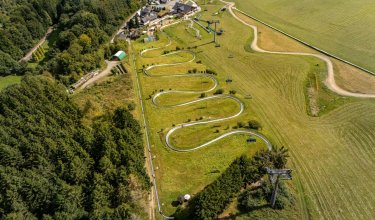 Luftaufnahme einer Sommerrodelbahn mit geschwungenen Bahnen neben Wald und Wiesen