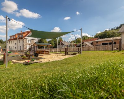 Spielplatz mit Sandkasten und grünem Sonnensegel, umgeben von Gras und Häusern im Hintergrund