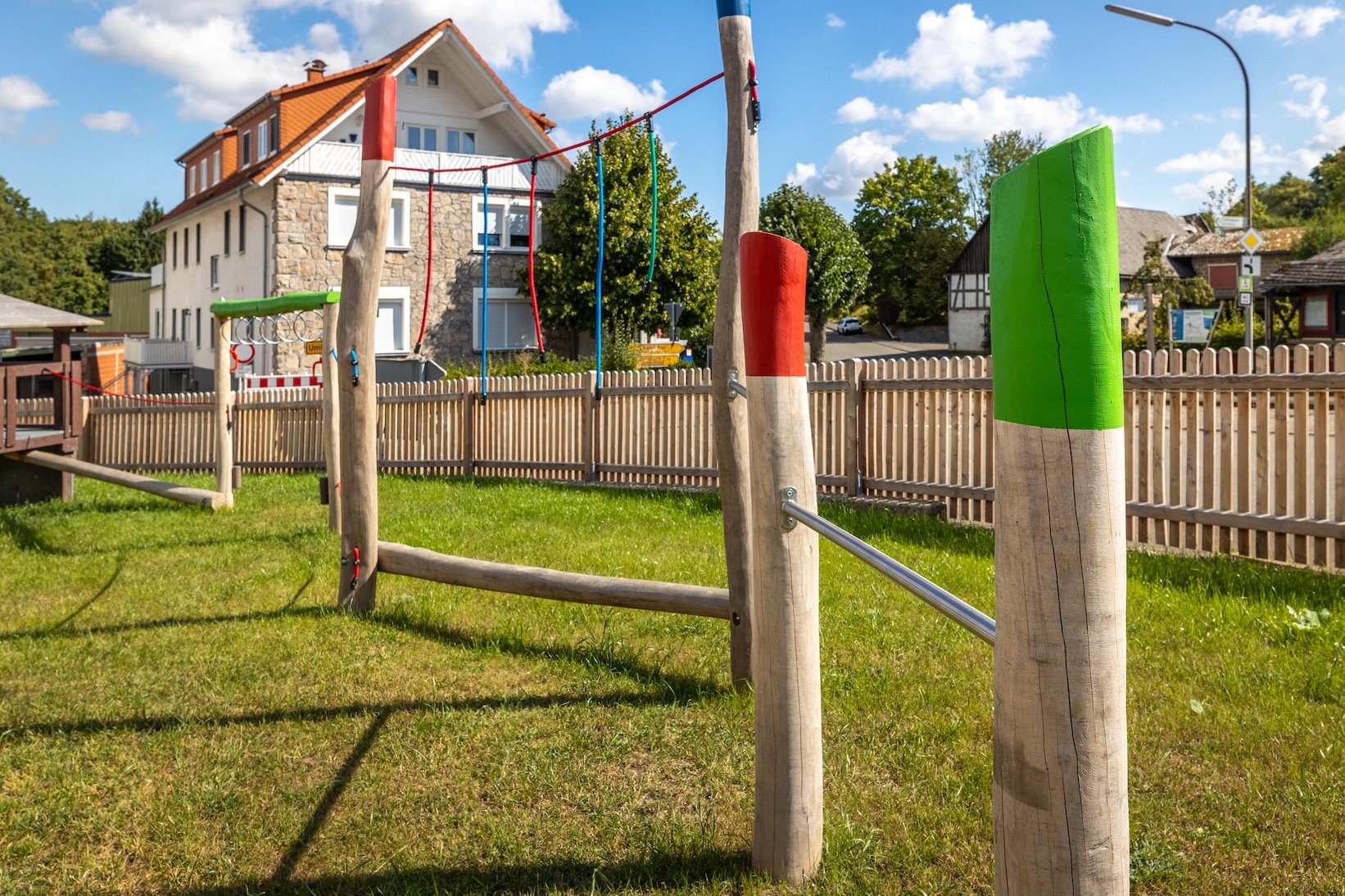 Holzklettergerüst mit bunten Seilen auf einem Spielplatz vor einem Zaun und Häusern