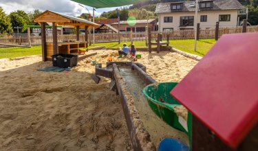 Sandkasten mit Wasserlauf und Spielhaus, Kinder spielen im Hintergrund, Haus und Zaun sichtbar