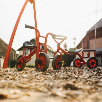 Zwei rote Kinderfahrzeuge auf Holzspänen vor einer Schaukel auf einem Spielplatz