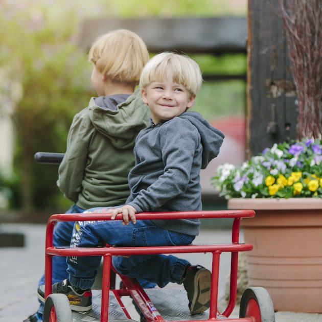 Zwei Kinder sitzen auf einem roten Dreirad, eines schaut lächelnd zurück, im Hintergrund Blumen