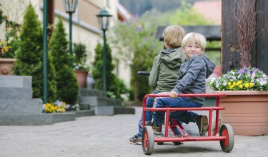Zwei Kinder sitzen auf einem roten Dreirad auf einem gepflasterten Weg neben Blumen und Laternen