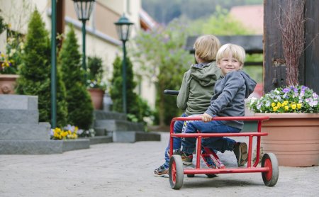 Zwei Kinder sitzen auf einem roten Dreirad auf einem gepflasterten Weg neben Blumen und Laternen