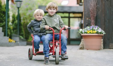 Zwei Jungen fahren zusammen auf einem roten Dreirad auf einem Hof mit Blumen im Hintergrund.