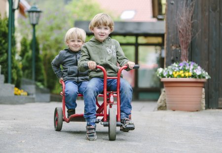 Zwei Jungen fahren zusammen auf einem roten Dreirad auf einem Hof mit Blumen im Hintergrund.