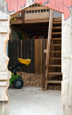 Blick durch eine Holztür auf eine Holztreppe und einen kleinen grünen Spielbagger mit gelber Schaufel