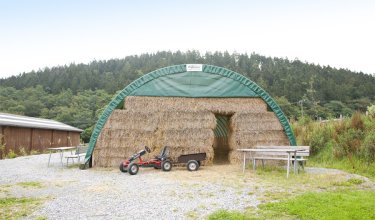 Grünes Zelt mit Heuballen und einem roten Kindertraktor davor, umgeben von Bäumen und Wiesen