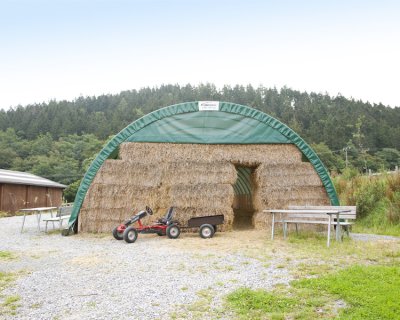 Grünes Zelt mit Heuballen und einem roten Kindertraktor davor, umgeben von Bäumen und Wiesen