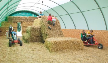 Kinder spielen in einer überdachten Scheune mit Heuballen und Tretfahrzeugen auf Strohboden