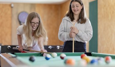 Zwei junge Frauen spielen lächelnd Billard an einem grünen Tisch in einem Raum mit orangefarbenen Wänden