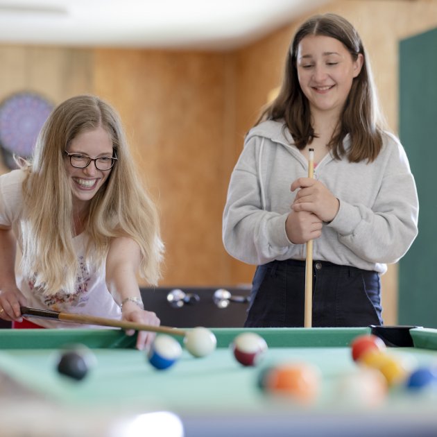 Zwei junge Frauen spielen lächelnd Billard an einem grünen Tisch in einem Raum mit orangefarbenen Wänden