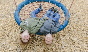 Zwei Kinder liegen kopfüber auf einer blauen Nestschaukel über Holzspänen am Boden.