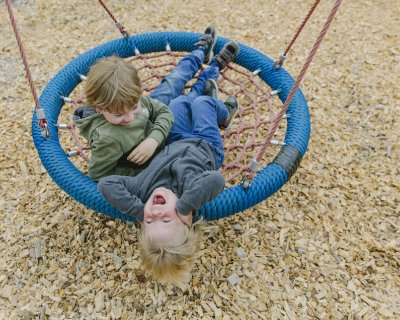 Zwei Kinder liegen lachend auf einer runden Schaukel über Holzspänen auf dem Spielplatz.