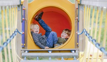 Zwei Kinder liegen lachend in einem gelben Spieltunnel auf einem Spielplatz
