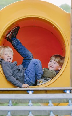 Zwei Kinder liegen lachend in einem gelben Spieltunnel auf einem Spielplatz
