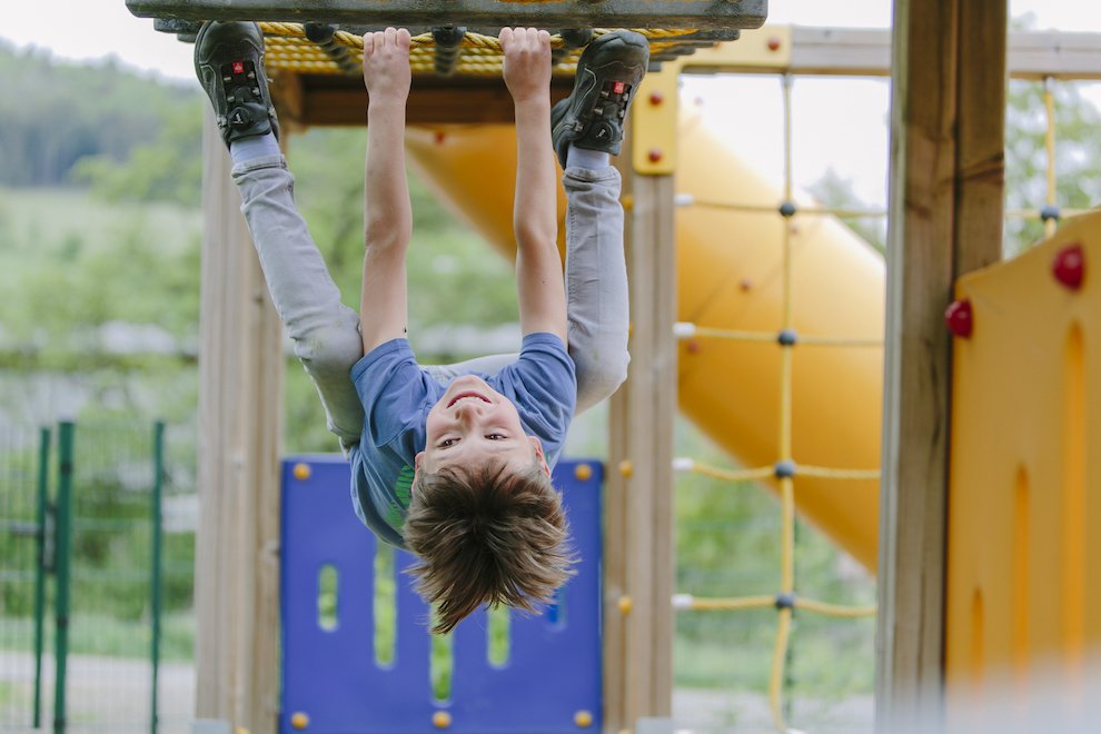 Junge hängt kopfüber an Klettergerüst auf Spielplatz mit Rutsche im Hintergrund
