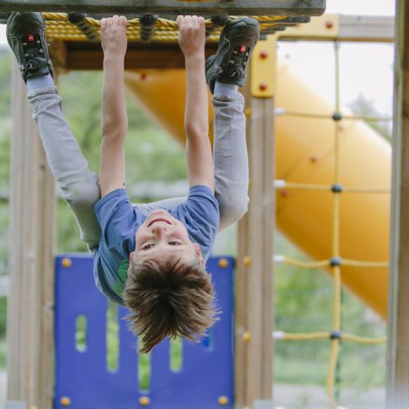 Junge hängt kopfüber an Klettergerüst auf Spielplatz mit Rutsche im Hintergrund
