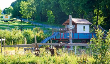 Spielplatz mit Rutsche und Holzhaus neben einem Zaun in einer grünen, bewaldeten Landschaft