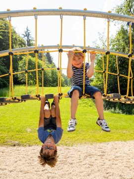 Zwei Kinder spielen an einem Klettergerüst im Freien, eines hängt kopfüber, das andere sitzt oben