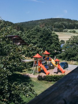 Blick auf einen Spielplatz mit roten Rutschen und grünen Bäumen vor bewaldetem Hügel unter blauem Himmel