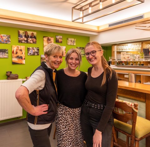 Drei lächelnde Frauen stehen nebeneinander in einem gemütlichen Raum mit grüner Wand und Bar im Hintergrund.