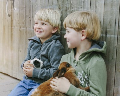 Zwei Jungen sitzen an einer Holzwand, jeder hält ein Kaninchen im Arm und lächelt