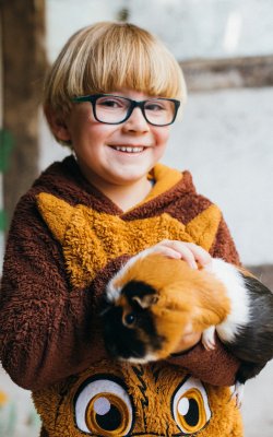 Lächelndes Kind mit Brille hält ein Meerschweinchen in den Armen, stehend vor einer Wand