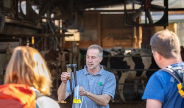 Mann erklärt zwei Geräte in einem Kuhstall, zwei Personen hören zu, Kühe im Hintergrund