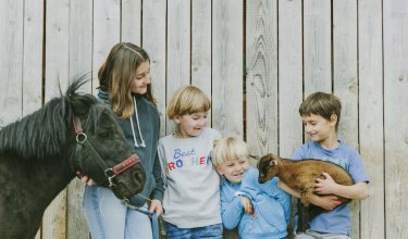 Vier Kinder vor einem Holzzaun, eines hält ein kleines braunes Zicklein, ein anderes führt ein schwarzes Pony an der Leine.